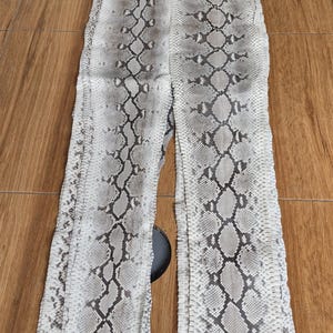 Op de afbeelding: Twee lange, smalle stukken slangenleer in een natuurlijk kleurenpalet van wit, grijs en bruin. Het slangenleer heeft een schubbenpatroon en ligt op een houten oppervlak. De stukken liggen naast elkaar, met een kleine opening ertussen.