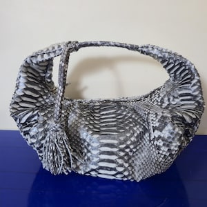 Könnte beinhalten: Eine graue und weiße Handtasche aus Schlangenhaut mit einem gebogenen Griff und einer Quaste. Die Tasche hat eine abgerundete Form und eine strukturierte Oberfläche mit einem Schuppenmuster. Der Griff ist ebenfalls aus Schlangenhaut.