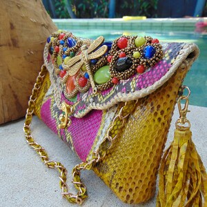 May include: A yellow, pink and green snakeskin clutch bag with a gold chain strap and a tassel. The bag has a beaded embellishment with a dragonfly design and colourful stones.