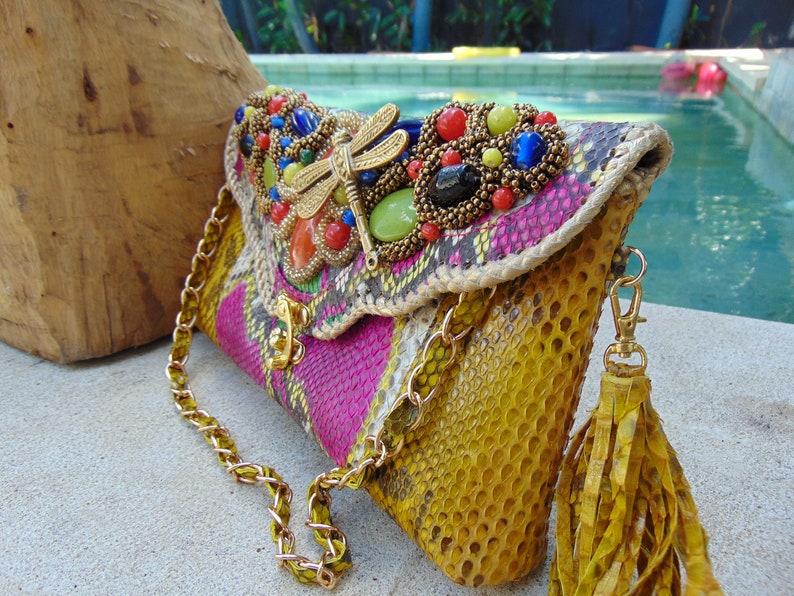 May include: A yellow, pink and green snakeskin clutch bag with a gold chain strap and a tassel. The bag has a beaded embellishment with a dragonfly design and colourful stones.