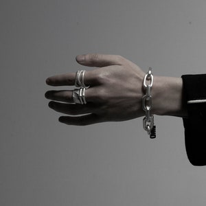 Puede incluir: Un brazalete de cadena de plata con un dise&ntilde;o de eslabones grandes. El brazalete se lleva en la mu&ntilde;eca de una persona. La persona tambi&eacute;n lleva varios anillos de plata en los dedos.