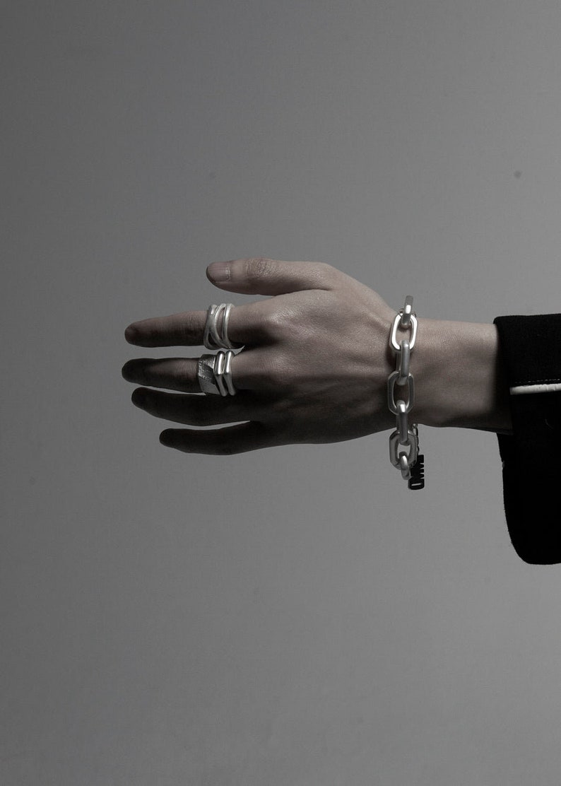 Puede incluir: Un brazalete de cadena de plata con un dise&ntilde;o de eslabones grandes. El brazalete se lleva en la mu&ntilde;eca de una persona. La persona tambi&eacute;n lleva varios anillos de plata en los dedos.
