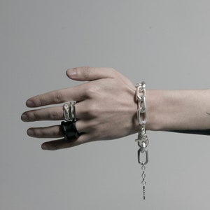 K&ouml;nnte beinhalten: Ein silbernes Kettenarmband mit einem gro&szlig;en Gliederdesign und einem silbernen Verschluss. Das Armband hat eine kleine silberne Kette, die vom Verschluss h&auml;ngt. Eine Person tr&auml;gt das Armband am Handgelenk. Die Person tr&auml;gt auch zwei silberne Ringe an ihren Fingern.