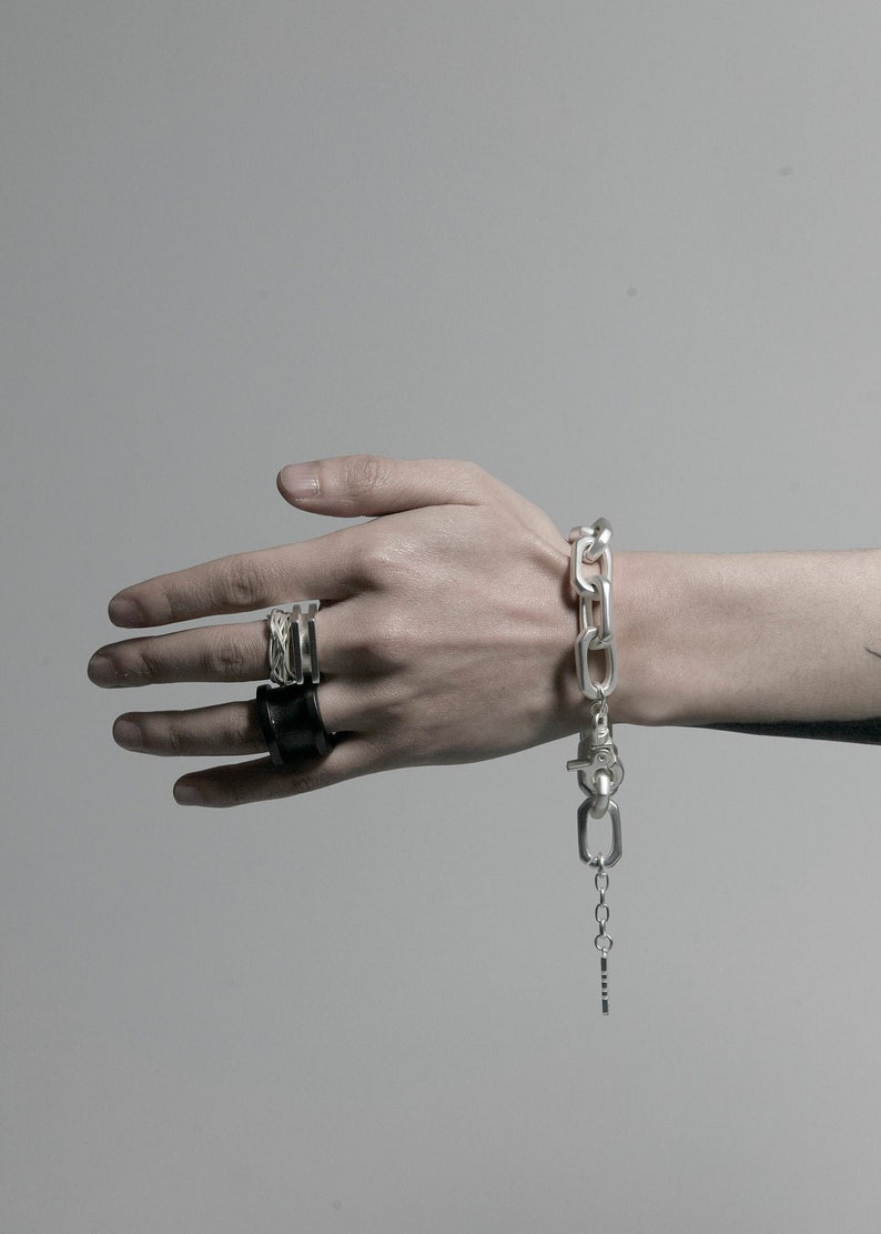 K&ouml;nnte beinhalten: Ein silbernes Kettenarmband mit einem gro&szlig;en Gliederdesign und einem silbernen Verschluss. Das Armband hat eine kleine silberne Kette, die vom Verschluss h&auml;ngt. Eine Person tr&auml;gt das Armband am Handgelenk. Die Person tr&auml;gt auch zwei silberne Ringe an ihren Fingern.