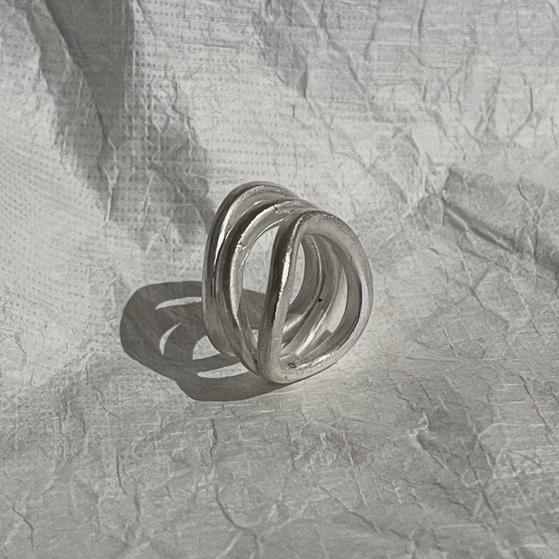 Puede incluir: Un anillo de plata con un dise&ntilde;o geom&eacute;trico &uacute;nico. El anillo est&aacute; hecho de m&uacute;ltiples bandas superpuestas que forman una forma triangular.