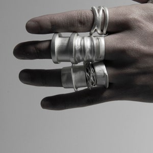 Puede incluir: Una mano con un conjunto de cinco anillos de plata. Los anillos est&aacute;n apilados en dos dedos y son de diferentes estilos y texturas.