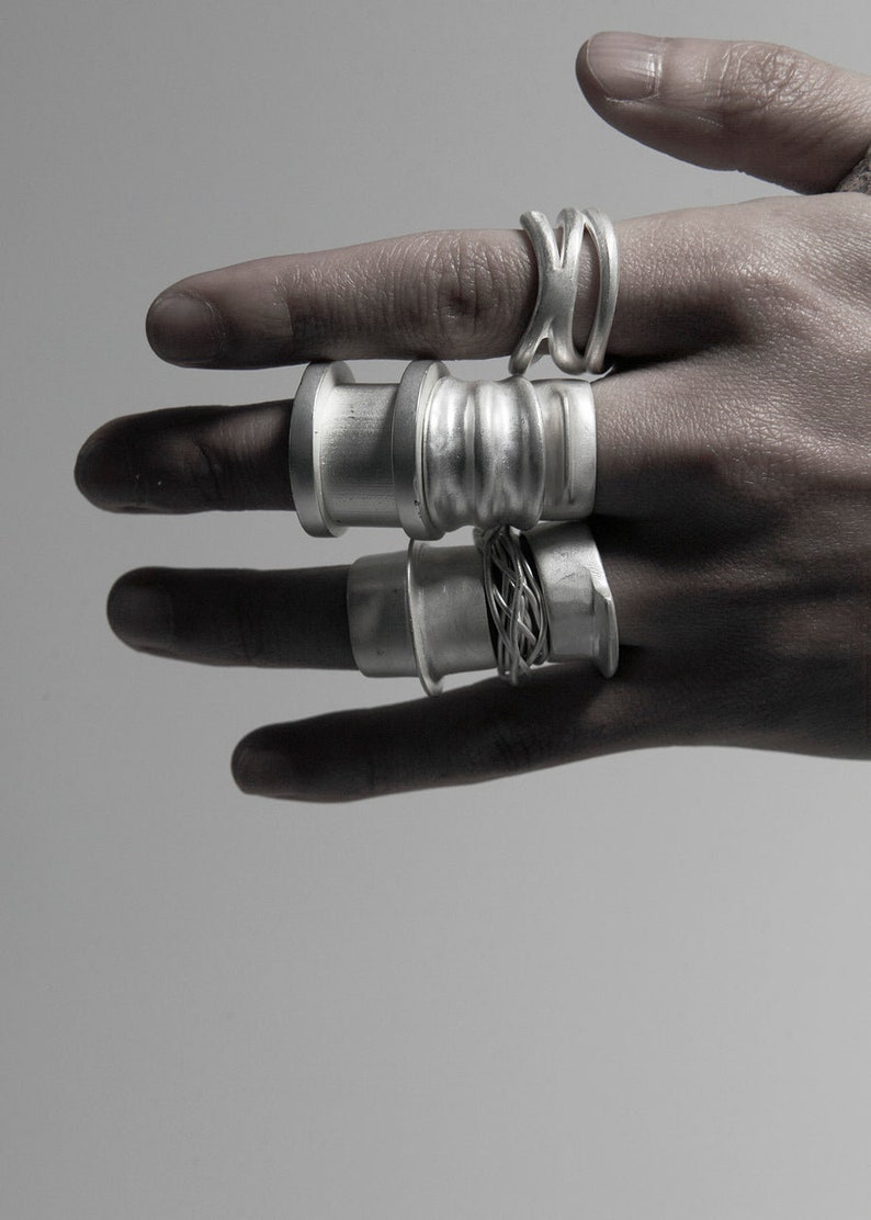 Puede incluir: Una mano con un conjunto de cinco anillos de plata. Los anillos est&aacute;n apilados en dos dedos y son de diferentes estilos y texturas.