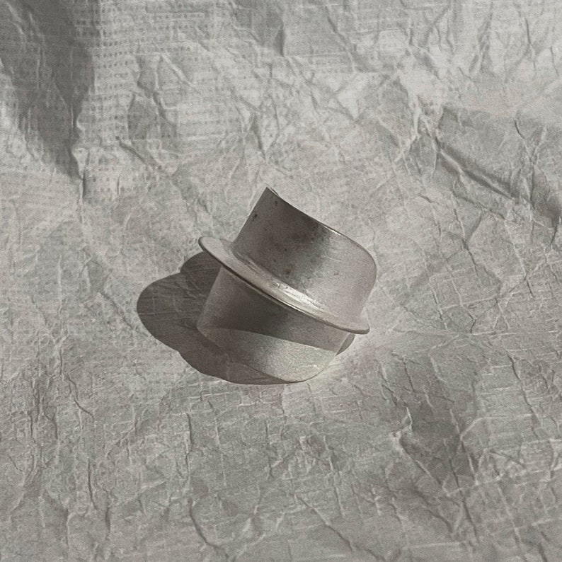 May include: A silver ring with a unique, abstract design. The ring is made of two pieces of metal that are joined together at an angle, creating a geometric shape.