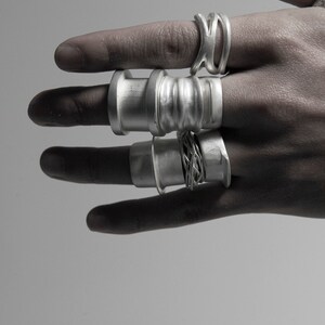 May include: A hand wearing five silver rings. The rings are all different styles, some are thick and some are thin. The rings are all made of metal and have a shiny finish.