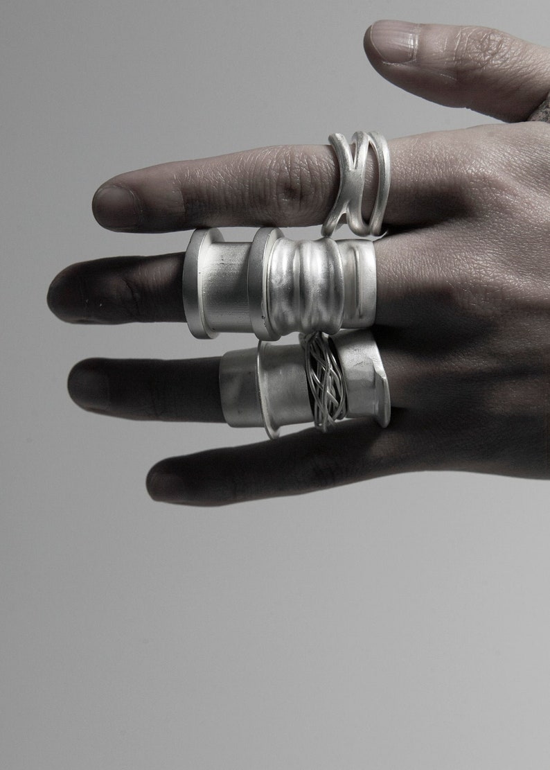 May include: A hand wearing five silver rings. The rings are all different styles, some are thick and some are thin. The rings are all made of metal and have a shiny finish.