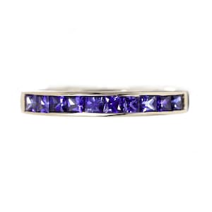 May include: A silver ring with a row of square, deep purple gemstones. The gemstones are set closely together, creating a continuous band of color. The ring is set against a white background.