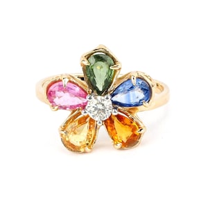 Op de afbeelding: Een gouden ring met een bloemmotief met een centrale diamant en vijf peervormige saffieren in verschillende kleuren: roze, groen, blauw, geel en oranje.