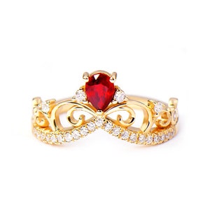 Puede incluir: Un anillo de oro con una piedra preciosa roja en el centro y un diseño de corona. El anillo está engastado con pequeños diamantes alrededor de la piedra preciosa y la corona.