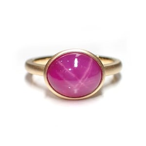 Könnte beinhalten: Ein Ring aus Gold mit einem rosa Stern-Saphir. Der Ring ist mit einem runden, cabochon geschliffenen Stern-Saphir mit einem sechsstrahligen Stern besetzt.