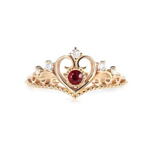 May include: A gold ring featuring a heart-shaped design with a red gemstone at its centre. The ring is adorned with small white gemstones.