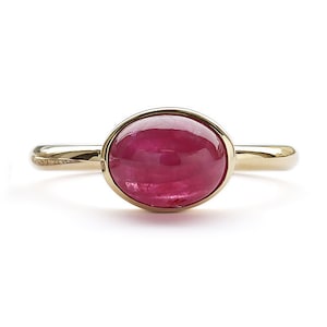 Puede incluir: Un anillo de oro con una piedra preciosa de rubí cabujón. El rubí es de color rojo intenso y tiene una superficie lisa y pulida.
