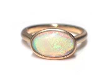 Könnte beinhalten: Ein goldener Ring mit einem ovalen Opal-Edelstein. Der Opal hat eine weiße Basis mit grün- und rosafarbenen, schillernden Blitzen.