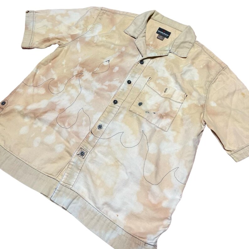 May include: A beige short-sleeved button-down shirt with a bleach-splattered design and black flame outlines. The shirt has a single chest pocket and a button-up collar.