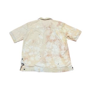 May include: A beige short-sleeved button-up shirt with a flame design embroidered on the back. The shirt has a faded, tie-dye effect.