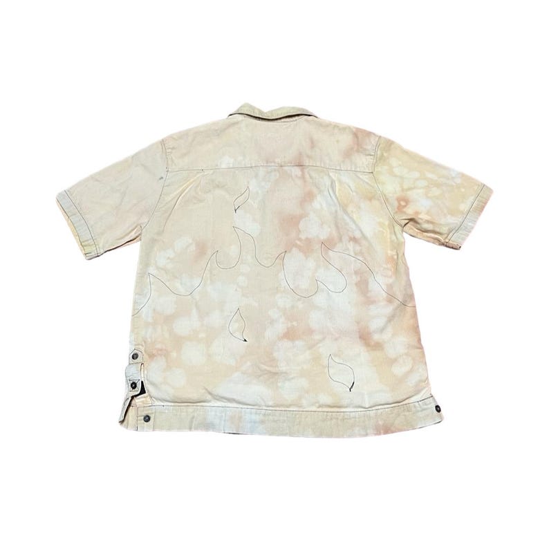 May include: A beige short-sleeved button-up shirt with a flame design embroidered on the back. The shirt has a faded, tie-dye effect.