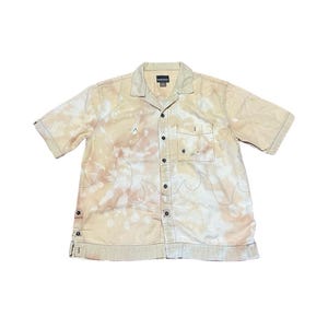 May include: A beige short-sleeved button-down shirt with a tie-dye pattern and a flame design. The shirt has a single chest pocket.