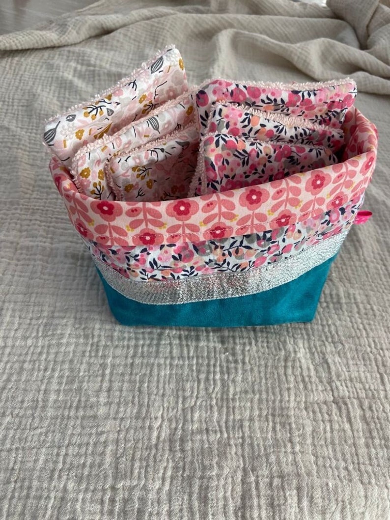 Puede incluir: Una cesta de almacenamiento de tela turquesa con ribete plateado y forro estampado floral rosa. La cesta est&aacute; llena de almohadillas de desmaquillaje reutilizables con estampado floral rosa y blanco.
