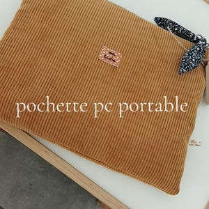Peut inclure: Housse d'ordinateur portable en velours côtelé moutarde avec le texte "pochette pc portable". La housse a une petite étiquette et un accent en tissu à motifs. La texture du velours côtelé est visible.