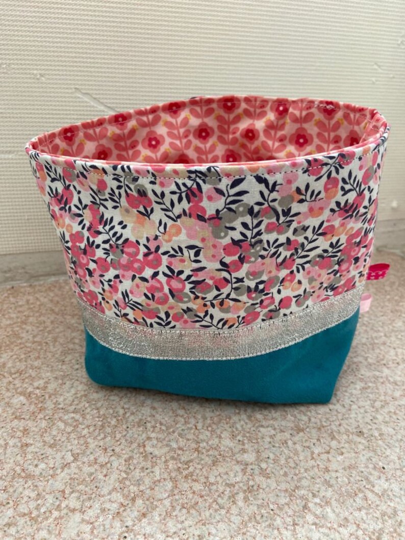 Puede incluir: Una cesta de almacenamiento de tela turquesa con un borde plateado y un forro con estampado floral. La cesta est&aacute; hecha de una tela suave y duradera y es perfecta para organizar art&iacute;culos peque&ntilde;os.