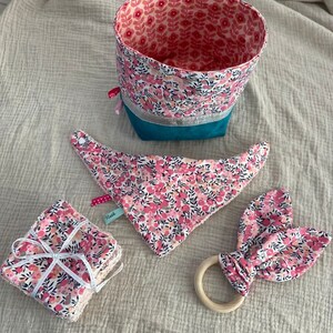 Puede incluir: Un conjunto de art&iacute;culos para beb&eacute;s con un estampado floral rosa, que incluye una cesta de almacenamiento de tela, un babero, un mordedor y toallitas lavables. La cesta tiene un forro turquesa y un ribete plateado. Las toallitas lavables est&aacute;n atadas con una cinta blanca.