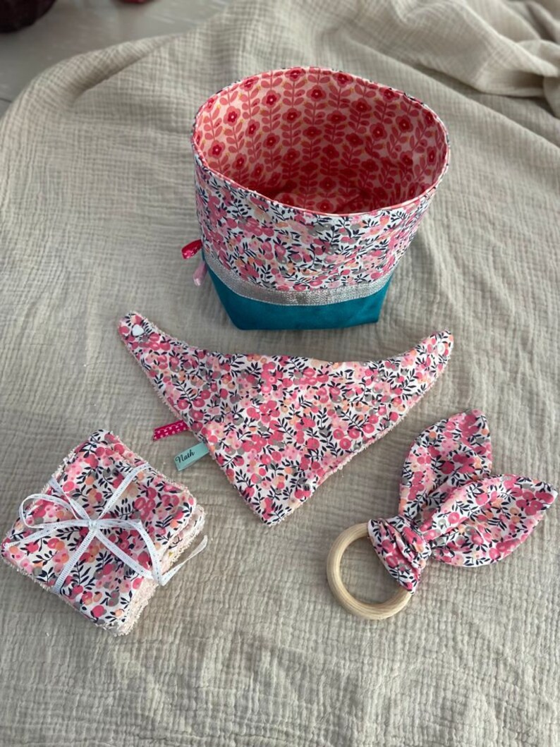 Puede incluir: Un conjunto de art&iacute;culos para beb&eacute;s con un estampado floral rosa, que incluye una cesta de almacenamiento de tela, un babero, un mordedor y toallitas lavables. La cesta tiene un forro turquesa y un ribete plateado. Las toallitas lavables est&aacute;n atadas con una cinta blanca.