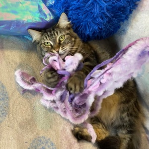 May include: A tabby cat with green eyes plays with a purple and white fluffy toy. The cat is lying on a light-colored blanket, with a blue fluffy pillow and iridescent fabric in the background.