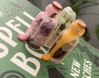 May include: Three small glass bottles with cork stoppers, each with a different colored wax seal. The bottles are filled with various crystals and herbs. The bottles are resting on a green book with the words "SPELL BOOK" and "NEW WITCHES" printed on the cover.