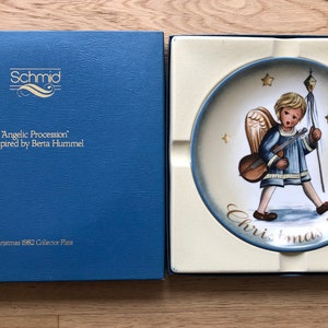 Può includere: Un piatto in ceramica blu e bianco con un design natalizio del 1982. Il piatto presenta un dipinto di un angelo che suona uno strumento musicale e il testo "Christmas 1982". Il piatto è in una scatola blu con il testo "Schmid" e "Angelic Procession" ispirato da Berta Hummel.