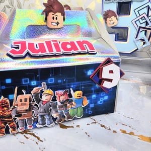 K&ouml;nnte beinhalten: Eine holografische Geschenkbox mit dem Namen "Julian" in roten Buchstaben. Die Box ist mit Roblox-Charakter-Ausschnitten und der Zahl 9 verziert. Im Hintergrund befindet sich eine gro&szlig;e blaue Zahl 6 mit Roblox-Charakteren.