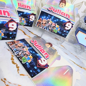 K&ouml;nnte beinhalten: Holografische Geburtstags-Gastgeschenkboxen mit dem Namen "Julian" und der Zahl "9". Die Boxen zeigen Roblox-Charaktere und eine Roblox-Charakterfigur oben. Die Boxen werden auf einer wei&szlig;en Oberfl&auml;che mit goldenen Akzenten pr&auml;sentiert.