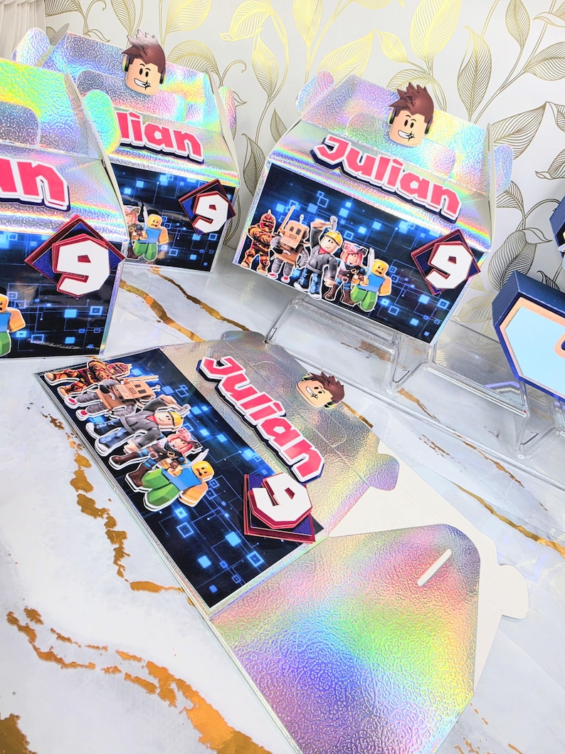 K&ouml;nnte beinhalten: Holografische Geburtstags-Gastgeschenkboxen mit dem Namen "Julian" und der Zahl "9". Die Boxen zeigen Roblox-Charaktere und eine Roblox-Charakterfigur oben. Die Boxen werden auf einer wei&szlig;en Oberfl&auml;che mit goldenen Akzenten pr&auml;sentiert.