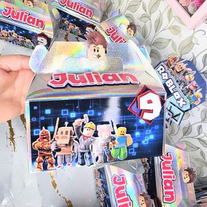 K&ouml;nnte beinhalten: Holografische Geschenkboxen mit dem Namen "Julian" in Rot und Wei&szlig;. Jede Box zeigt Roblox-Charaktere und eine "9" auf der Vorderseite. Ein Partygeschenk zum Thema Roblox.