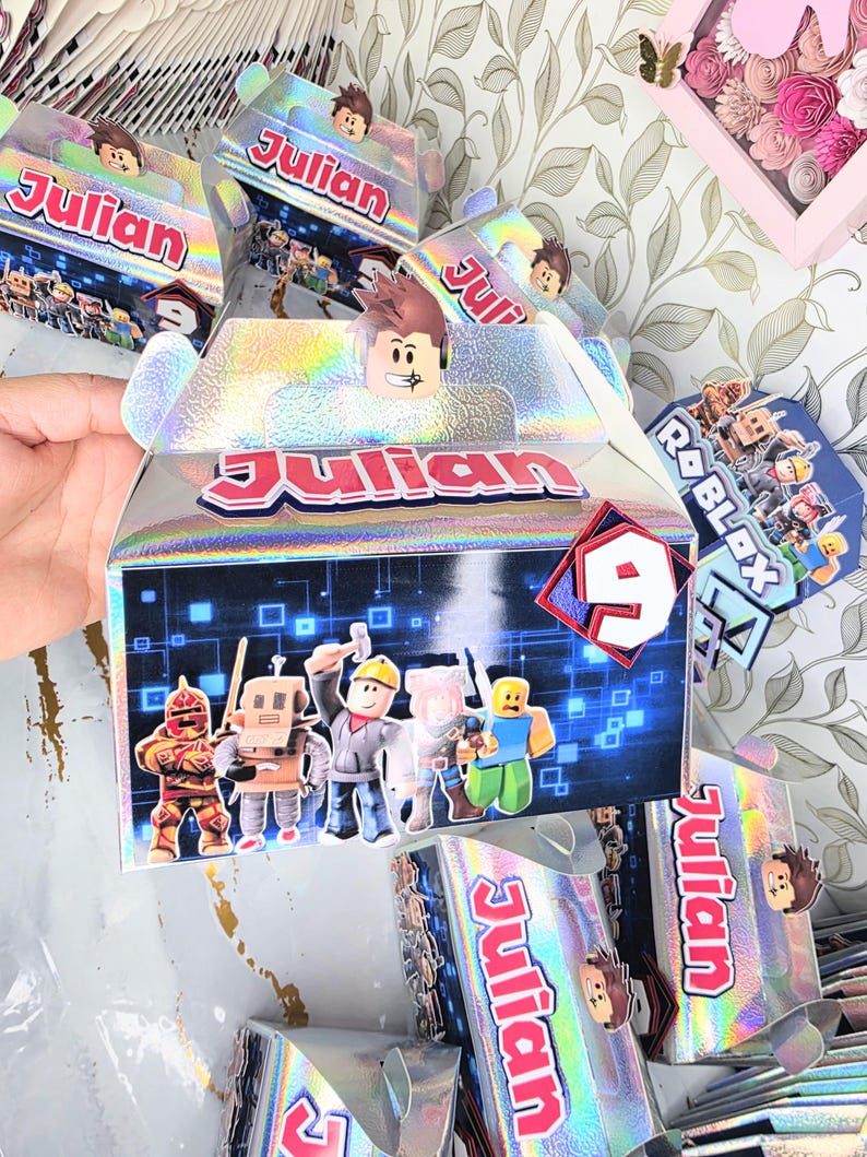 K&ouml;nnte beinhalten: Holografische Geschenkboxen mit dem Namen "Julian" in Rot und Wei&szlig;. Jede Box zeigt Roblox-Charaktere und eine "9" auf der Vorderseite. Ein Partygeschenk zum Thema Roblox.