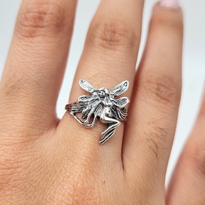 May include: A silver ring featuring a detailed fairy design with wings outstretched. The fairy is sitting on a branch with leaves.