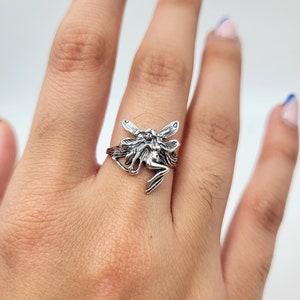 May include: A silver ring featuring a detailed fairy design with outstretched wings. The fairy is depicted in a sitting position with its wings spread out.
