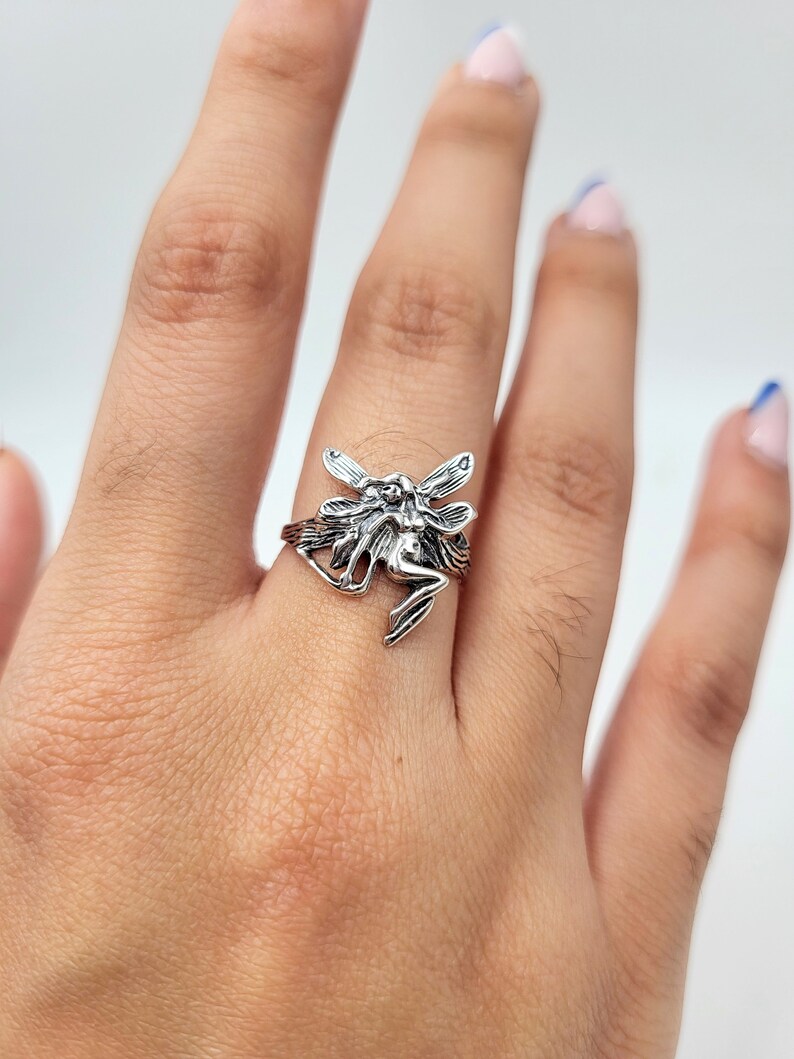 May include: A silver ring featuring a detailed fairy design with outstretched wings. The fairy is depicted in a sitting position with its wings spread out.