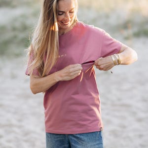 Puede incluir: Una camiseta rosa polvoriento con la palabra "mama" bordada en el pecho. La camiseta se combina con vaqueros de mezclilla azules. La modelo lleva pulseras doradas. La camisa tiene una abertura oculta.