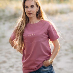Puede incluir: Camiseta rosa polvoriento con la palabra "mama" en letras doradas. La camiseta tiene cuello redondo y mangas cortas. La modelo lleva vaqueros azules y pulseras doradas.