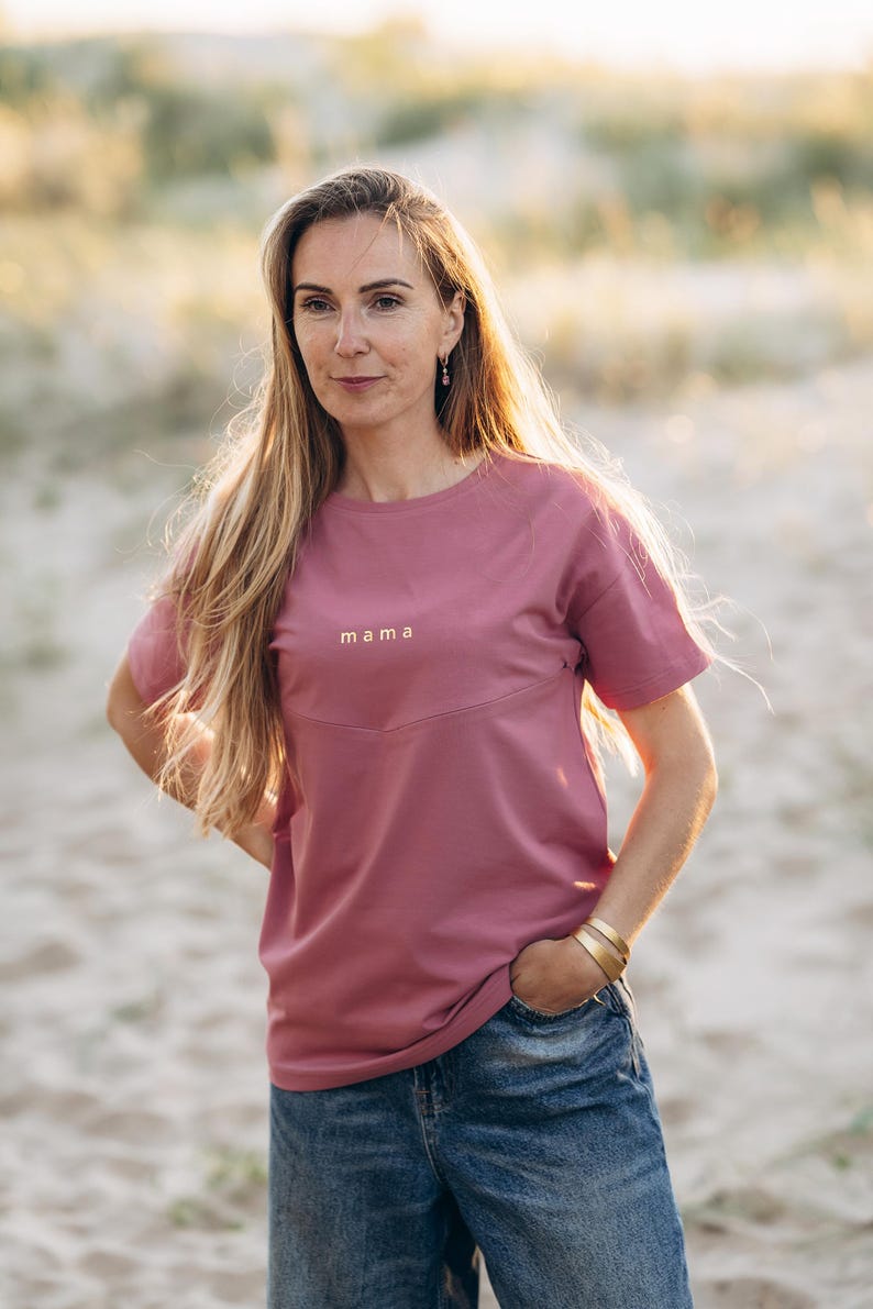 Puede incluir: Camiseta rosa polvoriento con la palabra "mama" en letras doradas. La camiseta tiene cuello redondo y mangas cortas. La modelo lleva vaqueros azules y pulseras doradas.