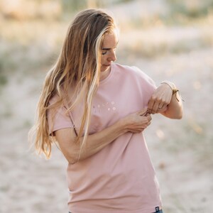 Puede incluir: Una camiseta rosa polvoriento con el texto "RELTO EL PO" impreso en la parte delantera. La camiseta se combina con vaqueros azules. La imagen est&aacute; tomada al aire libre con luz natural suave.