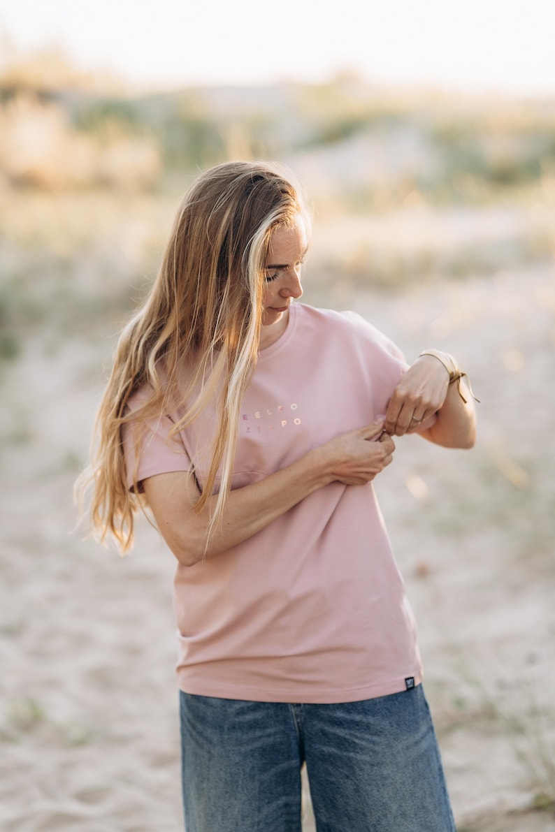Puede incluir: Una camiseta rosa polvoriento con el texto "RELTO EL PO" impreso en la parte delantera. La camiseta se combina con vaqueros azules. La imagen est&aacute; tomada al aire libre con luz natural suave.