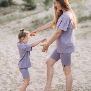 Puede incluir: Una mujer y una ni&ntilde;a con camisetas y pantalones cortos de ciclista lilas a juego en una playa de arena. La mujer est&aacute; agarrando la mano de la ni&ntilde;a. Los conjuntos tienen un dise&ntilde;o minimalista, adecuado para ropa informal.