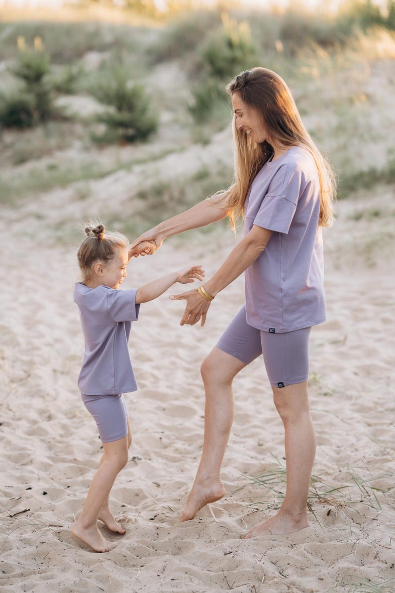 Puede incluir: Una mujer y una ni&ntilde;a con camisetas y pantalones cortos de ciclista lilas a juego en una playa de arena. La mujer est&aacute; agarrando la mano de la ni&ntilde;a. Los conjuntos tienen un dise&ntilde;o minimalista, adecuado para ropa informal.