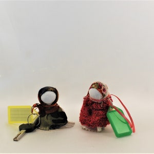 May include: Two small fabric dolls with embroidered faces and colorful clothing. The doll on the left is wearing a green and brown dress and a red scarf. The doll on the right is wearing a red dress with a floral print and a white head scarf. Both dolls are holding keychains.