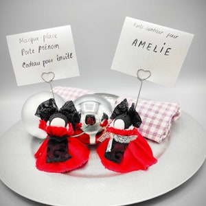 Peut inclure: Deux ornements de poupée rouge et noir avec des nœuds noirs et des visages blancs. Les poupées sont assises sur une assiette argentée avec une serviette à carreaux blancs et roses. Deux cartes de place sont attachées aux poupées avec des pinces en forme de cœur argentées. Les cartes de place indiquent "Marque-place Porte-prénom Cadeau pour invité" et "Porte bonheur pour AMELIE".